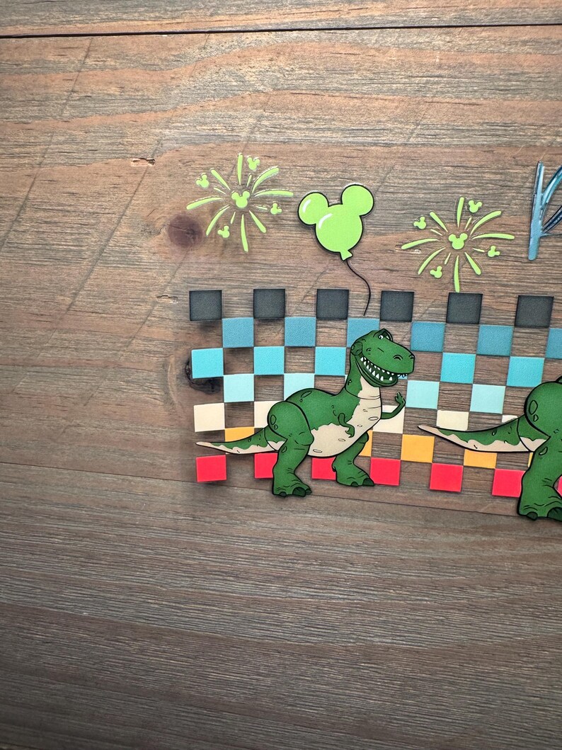 Puede incluir: Una manualidad de papel en capas que presenta un dinosaurio de dibujos animados verde frente a un patr&oacute;n de cuadros que cambia de negro a azul, beige, naranja y rojo. Sobre el dinosaurio hay fuegos artificiales verde claro y un globo con forma de Mickey.