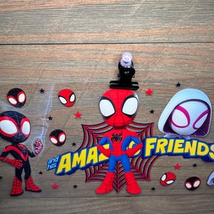 May include: A clear acrylic sign featuring cartoon characters from Spidey and His Amazing Friends, including Spider-Man, Ghost-Spider, and Miles Morales, with yellow and blue text, red spider web graphics, and small black and red stars.