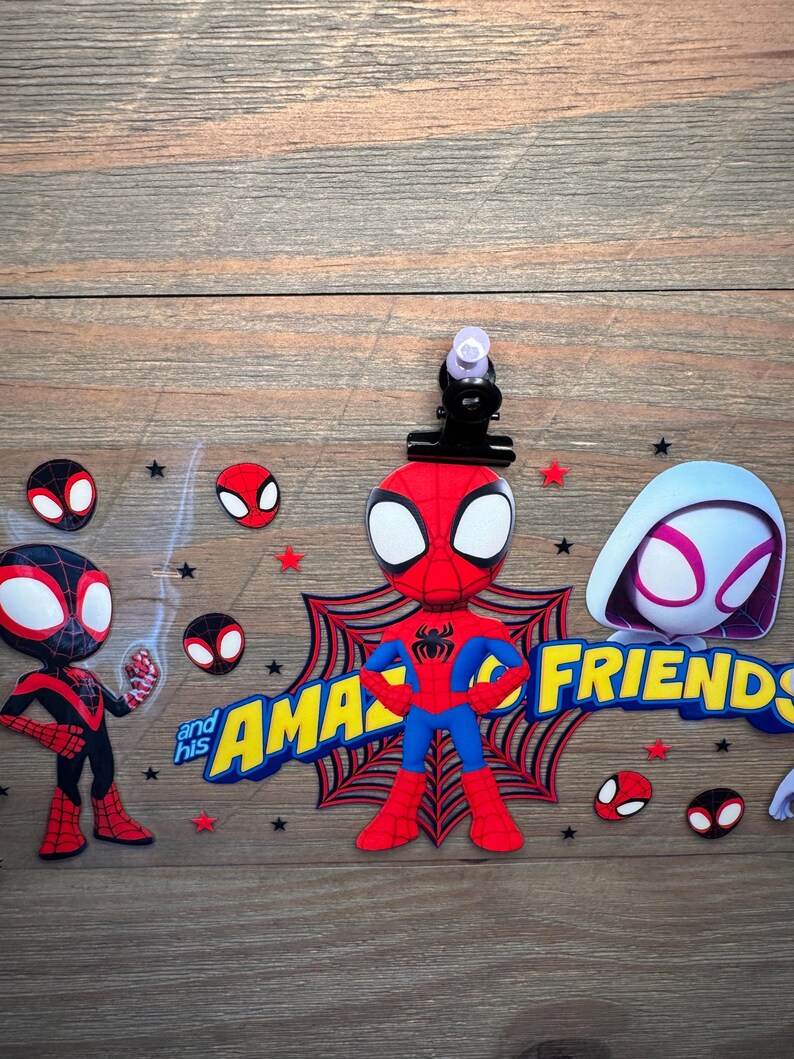May include: A clear acrylic sign featuring cartoon characters from Spidey and His Amazing Friends, including Spider-Man, Ghost-Spider, and Miles Morales, with yellow and blue text, red spider web graphics, and small black and red stars.