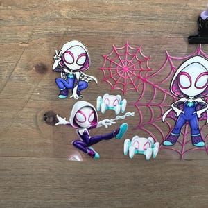 May include: A collection of Spider-Gwen themed stickers and embellishments. The stickers feature the character in various poses, with a white and pink color scheme. Also included are pink spiderwebs and small white spiders.