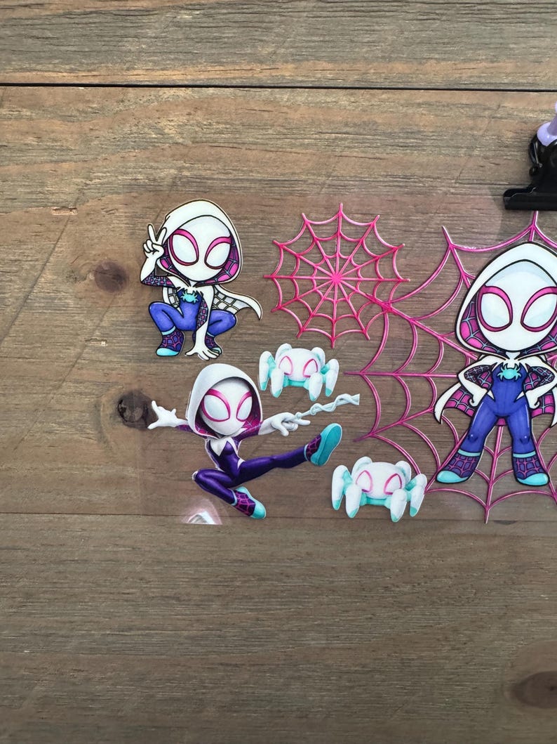May include: A collection of Spider-Gwen themed stickers and embellishments. The stickers feature the character in various poses, with a white and pink color scheme. Also included are pink spiderwebs and small white spiders.