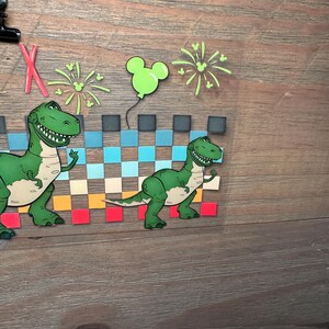 Puede incluir: Una hoja de transferencia transparente con dos dinosaurios verdes de dibujos animados, un patr&oacute;n de cuadros en azul, beige y rojo, fuegos artificiales de color verde claro y un globo verde claro con forma de Mickey Mouse. Incluye una letra X roja.