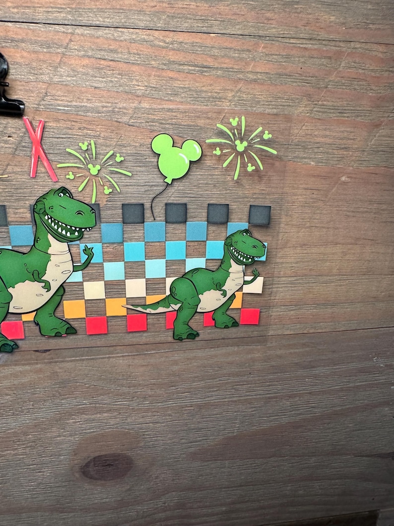 Puede incluir: Una hoja de transferencia transparente con dos dinosaurios verdes de dibujos animados, un patr&oacute;n de cuadros en azul, beige y rojo, fuegos artificiales de color verde claro y un globo verde claro con forma de Mickey Mouse. Incluye una letra X roja.