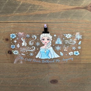 May include: A clear plastic luggage tag featuring a cartoon illustration of a woman with a blue dress and white hair. The tag includes snowflakes, flowers, and the text "The Cold Never Bothered Me Anyway."