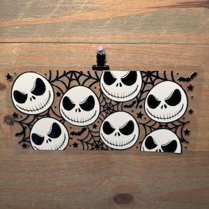 May include: A clear, rectangular sticker featuring eight Jack Skellington faces in a repeating pattern. The design includes a black spiderweb, bats, and stars. The sticker is held in place by a black clip with a purple push pin.