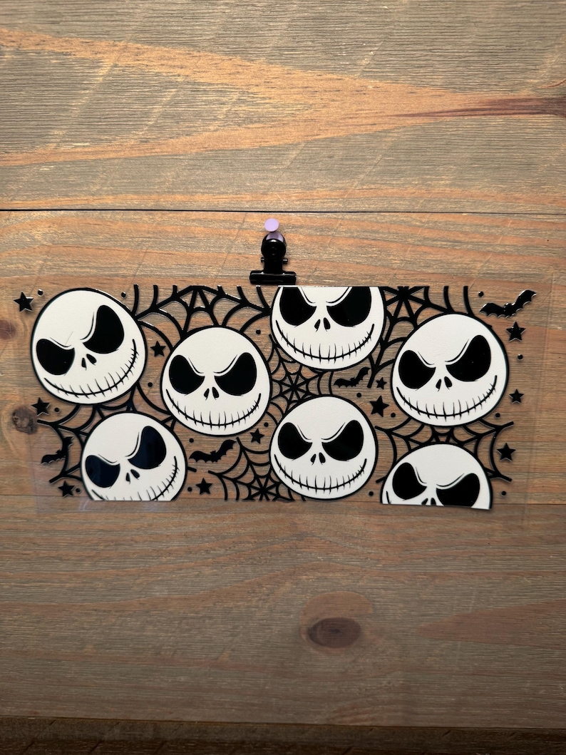 May include: A clear, rectangular sticker featuring eight Jack Skellington faces in a repeating pattern. The design includes a black spiderweb, bats, and stars. The sticker is held in place by a black clip with a purple push pin.