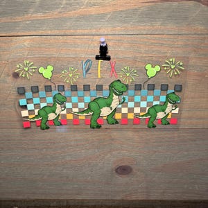 Puede incluir: Una calcoman&iacute;a rectangular transparente con tres dinosaurios T-Rex verdes frente a un patr&oacute;n de cuadros horizontal en tonos azul, amarillo y rojo. El dise&ntilde;o incluye fuegos artificiales verde claro, dos globos con forma de Mickey y el texto REX.