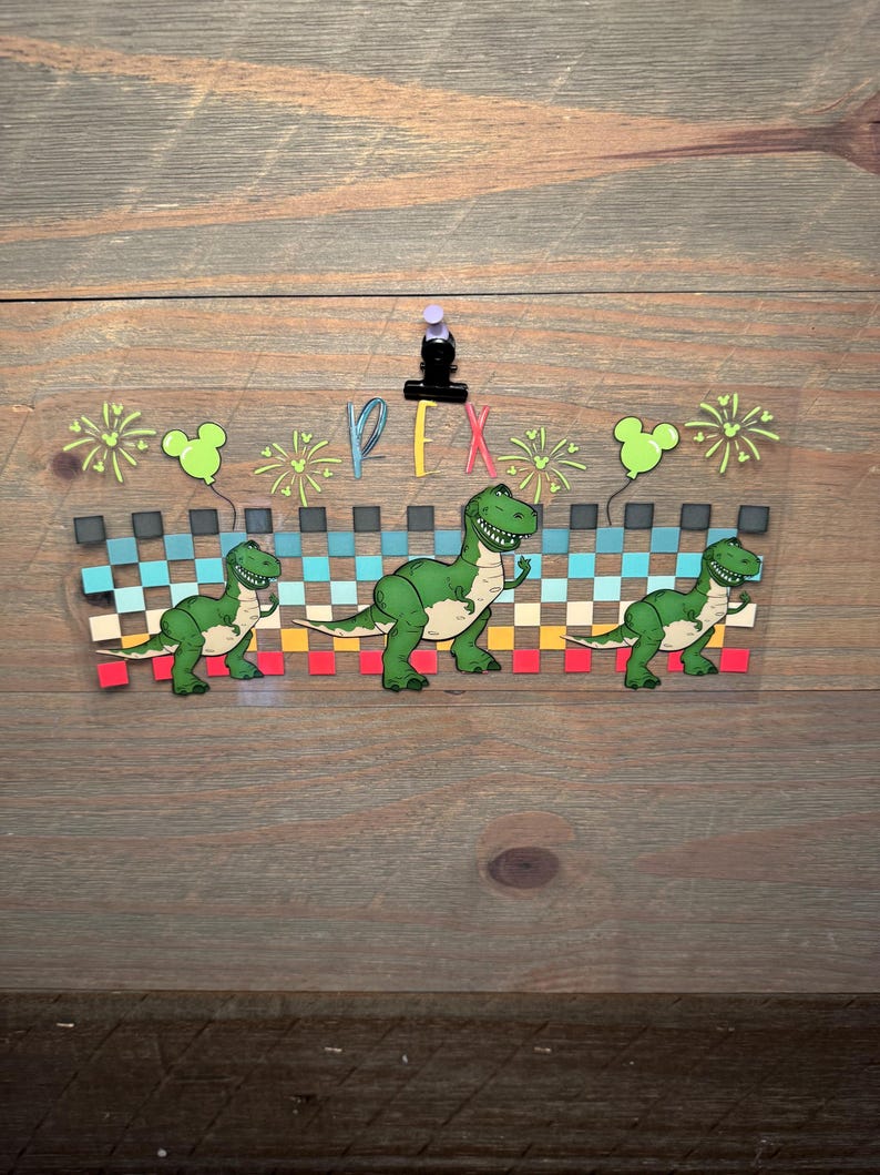 Puede incluir: Una calcoman&iacute;a rectangular transparente con tres dinosaurios T-Rex verdes frente a un patr&oacute;n de cuadros horizontal en tonos azul, amarillo y rojo. El dise&ntilde;o incluye fuegos artificiales verde claro, dos globos con forma de Mickey y el texto REX.