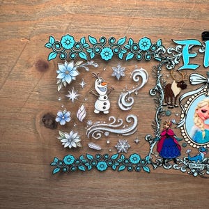 Puede incluir: Una l&aacute;mina de pegatinas transparente con personajes y elementos de la pel&iacute;cula "Frozen". El dise&ntilde;o incluye im&aacute;genes de Olaf, Elsa, Anna, copos de nieve y detalles florales en tonos azules y blancos. El texto "Frozen" es visible.