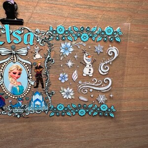 Puede incluir: Una l&aacute;mina de pegatinas transparente con Elsa de Frozen. El dise&ntilde;o incluye un retrato de Elsa, Olaf el mu&ntilde;eco de nieve y otros personajes, junto con detalles florales y de copos de nieve en tonos azules y blancos. El texto "Elsa" y "Frozen" tambi&eacute;n es visible.
