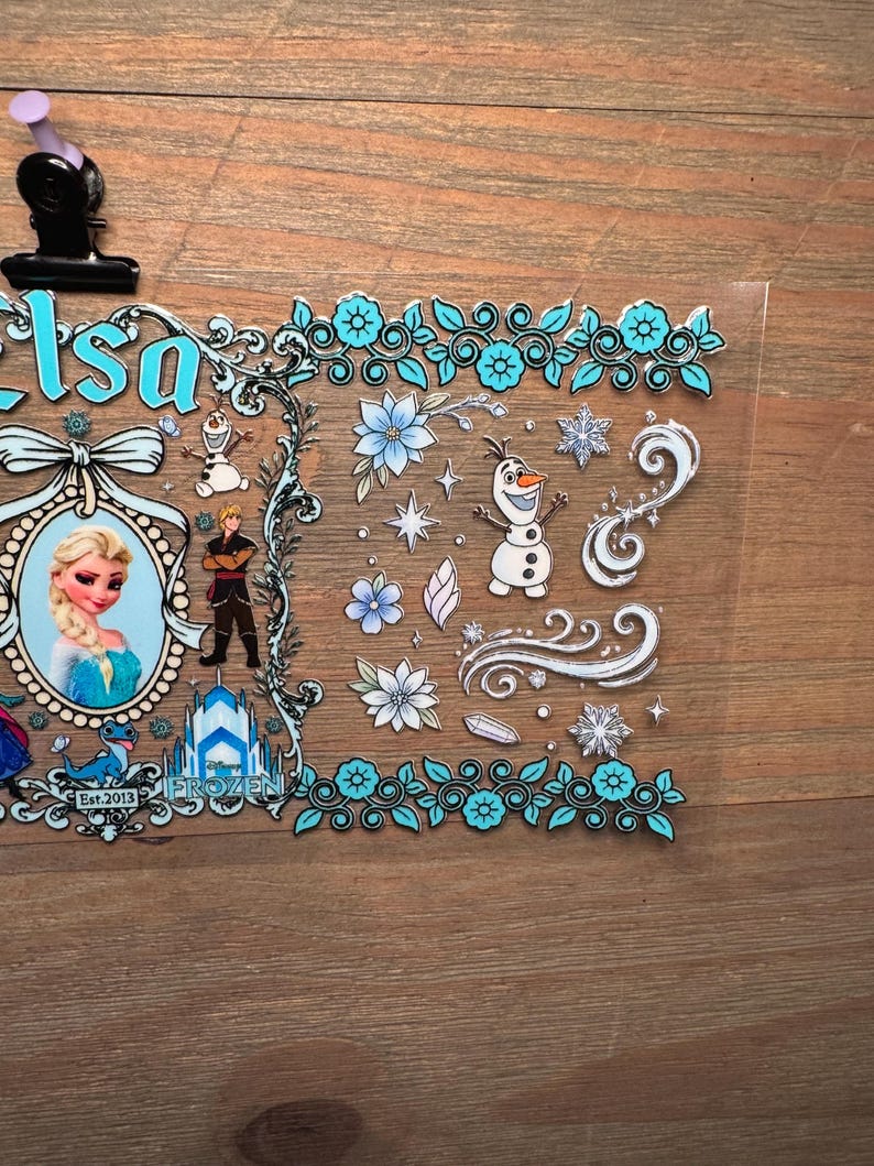 Puede incluir: Una l&aacute;mina de pegatinas transparente con Elsa de Frozen. El dise&ntilde;o incluye un retrato de Elsa, Olaf el mu&ntilde;eco de nieve y otros personajes, junto con detalles florales y de copos de nieve en tonos azules y blancos. El texto "Elsa" y "Frozen" tambi&eacute;n es visible.