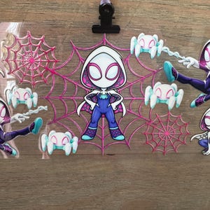 May include: A clear plastic sheet featuring Spider-Gwen character stickers. The stickers include a central Spider-Gwen figure, spider webs, and small spider figures in shades of pink, white, and blue. A black clip and purple push pin are at the top.