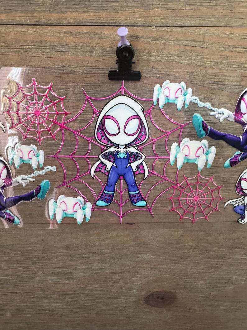 May include: A clear plastic sheet featuring Spider-Gwen character stickers. The stickers include a central Spider-Gwen figure, spider webs, and small spider figures in shades of pink, white, and blue. A black clip and purple push pin are at the top.