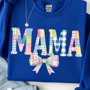 Peut inclure: Un sweat-shirt bleu roi avec le mot "MAMA" en lettres patchwork et un nœud papillon patchwork. Les lettres et le nœud présentent un mélange de motifs à carreaux pastel. Un collier argenté et un jean délavé clair sont également dans l'image.