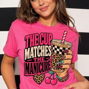 Op de afbeelding: Een felroze t-shirt met korte mouwen en een afbeelding van een hand die een beker met een ruitjes- en luipaardpatroon vasthoudt. De tekst luidt THE CUP MATCHES THE MANICURE, vergezeld van een hart met luipaardprint en twee rode kersen.