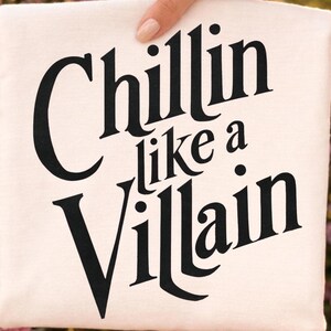 Puede incluir: Una camiseta de color crema con el texto negro "Chillin like a Villain" impreso en una fuente serif audaz y estilizada. El dise&ntilde;o est&aacute; centrado en la parte delantera de la prenda, con las palabras dispuestas en tres l&iacute;neas distintas sobre la tela.