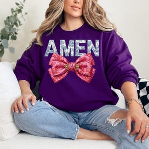 Op de afbeelding: Een paarse sweatshirt met het woord "AMEN" in witte letters met een blauw bloemenpatroon. Een grote roze strik met een gouden sluiting is onder het woord gecentreerd. De strik heeft een bloemenpatroon.