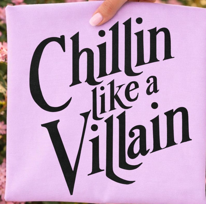 Puede incluir: Una camiseta de color morado claro con el texto negro "Chillin like a Villain" impreso en una fuente serif negrita y estilizada. Las letras est&aacute;n dispuestas en tres l&iacute;neas, con la palabra "Chillin" arriba, "like a" en el medio y "Villain" abajo.