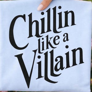 Puede incluir: Una camiseta azul claro con el texto negro "Chillin like a Villain" impreso en una fuente serif audaz y estilizada en el centro. Las letras est&aacute;n dispuestas en tres l&iacute;neas con una ligera orientaci&oacute;n diagonal para las palabras superior e inferior.
