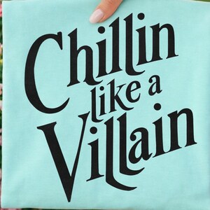 Puede incluir: Una camiseta de color turquesa claro con el texto negro "Chillin like a Villain" impreso en una fuente serif negrita y estilizada. Las letras est&aacute;n dispuestas en tres l&iacute;neas con la palabra "Chillin" arriba, "like a" en el medio y "Villain" abajo.