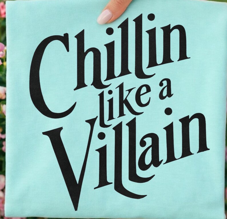 Puede incluir: Una camiseta de color turquesa claro con el texto negro "Chillin like a Villain" impreso en una fuente serif negrita y estilizada. Las letras est&aacute;n dispuestas en tres l&iacute;neas con la palabra "Chillin" arriba, "like a" en el medio y "Villain" abajo.