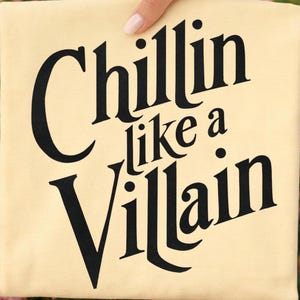 Puede incluir: Una camiseta de color crema claro con texto negro en negrita que dice "Chillin like a Villain" en una fuente serif estilizada. Las letras est&aacute;n centradas en la tela, con la palabra "Chillin" en la parte superior, "like a" en el medio y "Villain" debajo.