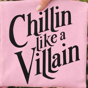 Puede incluir: Una camiseta rosa claro con el texto negro "Chillin like a Villain" impreso en una fuente serif audaz y estilizada. Las letras est&aacute;n dispuestas en tres l&iacute;neas, con la palabra "Chillin" en la parte superior, "like a" en el medio y "Villain" abajo.