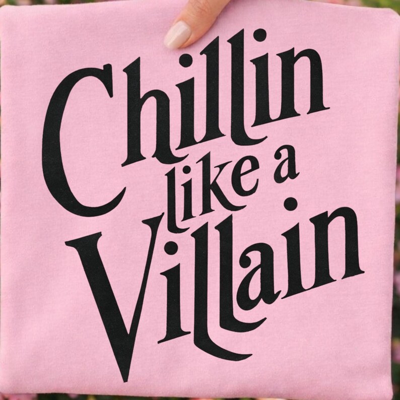 Puede incluir: Una camiseta rosa claro con el texto negro "Chillin like a Villain" impreso en una fuente serif audaz y estilizada. Las letras est&aacute;n dispuestas en tres l&iacute;neas, con la palabra "Chillin" en la parte superior, "like a" en el medio y "Villain" abajo.