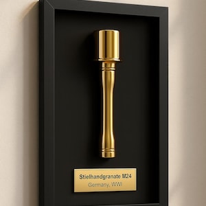 Könnte beinhalten: Gerahmte Präsentation einer goldfarbenen Stielhandgranate M24, einer deutschen Handgranate aus dem Ersten Weltkrieg. Die Granate ist auf schwarzem Hintergrund in einem schwarzen Rahmen montiert. Eine goldene Plakette darunter lautet "Stielhandgranate M24 Germany, WWI."