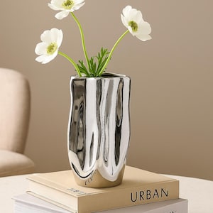 Może przedstawiać: Srebrny wazon o organicznym kształcie, z białymi kwiatami anemonów z zielonymi łodygami. Wazon stoi na dwóch książkach, z których jedna ma napis "URBAN", a druga "THE FASHION". Wazon ma metaliczne wykończenie i unikalny design.