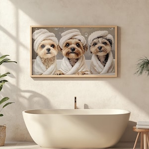 Op de afbeelding: Drie honden die witte badjassen en handdoeken op hun hoofd dragen, kijken naar de camera. De honden zijn in een ingelijst beeld met een lichtbruine houten lijst.