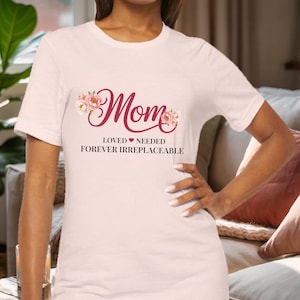 May include: A light pink t-shirt with the word "Mom" in a cursive, burgundy font, adorned with floral accents. Below, the text reads "LOVED NEEDED FOREVER IRREPLACEABLE" in a simple, sans-serif font. The shirt is short-sleeved.