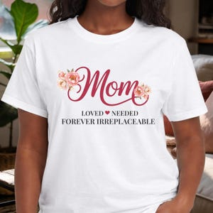 May include: White t-shirt with the word "Mom" in a cursive, burgundy font, adorned with floral accents. Below, the text reads "LOVED NEEDED FOREVER IRREPLACEABLE" in a simple, black font. The shirt is a classic crew neck.