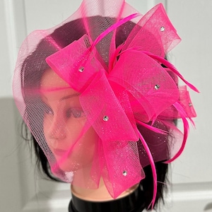 May include: A vibrant pink fascinator with a net veil and bow design. The headpiece features a large bow with rhinestone accents and pink feathers. The fascinator is displayed on a mannequin head.