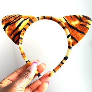 May include: A tiger ear headband with orange, black, and yellow stripes. The headband is made of a soft, plush material and features two upright ears. The design mimics a tiger's fur pattern, suitable for costumes or themed events.