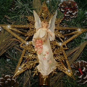 May include: A gold glitter star Christmas tree topper featuring an angel in a white gown holding a horn and a basket of pink roses. The angel has white wings and is set against a backdrop of pine needles and pine cones.