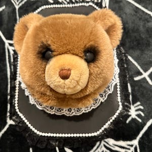 以下が含まれることがあります： 黒いプレートに取り付けられた茶色のテディベアのぬいぐるみヘッド。クマには暗い色の目と茶色の鼻があります。白いレースの襟と黒いフリルの縁取りがクマの首元を囲んでおり、黒いプレートの中央に取り付けられています。