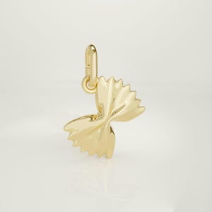 Könnte beinhalten: Ein goldfarbener Anhänger in Form von Farfalle-Nudeln. Der Anhänger hat oben eine Schlaufe zum Befestigen an einer Halskette oder einem Armband. Die Nudelform ist mit gerüschten Kanten und einem Schleifendesign versehen.