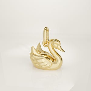 Può includere: Un ciondolo in metallo color oro lucido a forma di cigno. Il cigno è raffigurato con le ali leggermente sollevate e il collo elegantemente curvato. Un piccolo anellino di giunzione di forma ovale è fissato sul dorso del cigno per appenderlo.