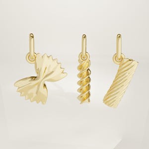 May include: Three gold-colored pasta-shaped charms. The charms include a bow tie, a fusilli, and a rigatoni. Each charm has a small loop at the top for hanging. The charms are displayed against a light background.