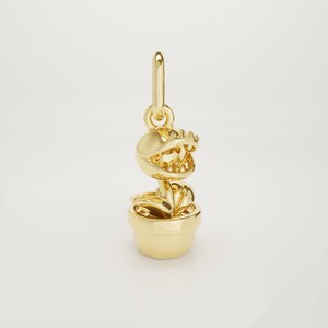 Op de afbeelding: Een goudkleurige bedel in de vorm van een cartoonplant met een grote, getande mond, geplaatst in een pot. De bedel heeft een lus aan de bovenkant om aan een ketting of armband te bevestigen. De bladeren en de pot van de plant zijn gedetailleerd.
