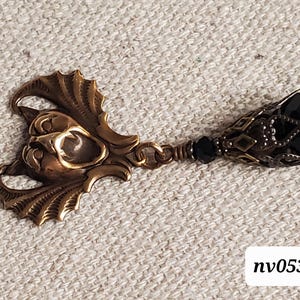 May include: A bronze-toned pendant with a detailed, stylized face and wing-like elements. The pendant is connected to a chain with black beads and a black, faceted teardrop-shaped bead. The text "nv053" is in the bottom right corner.