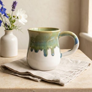 May include: A handmade ceramic mug with a white base and a green and blue glaze that drips down the sides. The mug sits on a beige linen napkin. A small white vase with blue and white flowers is in the background.
