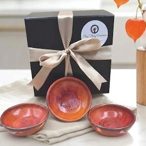 Può includere: Tre piccole ciotole rotonde in ceramica con una glassa lucida, screziata di rosso e arancione. Sono appoggiate su un panno color neutro davanti a una scatola regalo nera legata con un nastro beige. La scatola ha un adesivo circolare con la scritta "Clay Tory Ceramics".