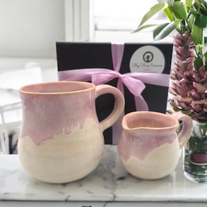 Puede incluir: Dos tazas de cerámica hechas a mano, una grande y otra pequeña, en una combinación de colores rosa y crema. Las tazas tienen forma redondeada con asas. Una caja de regalo negra con una cinta rosa en el fondo.