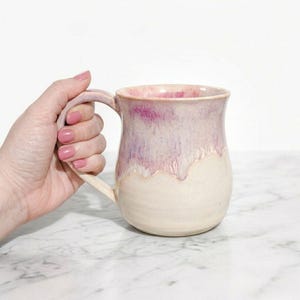 Peut inclure: Une tasse en céramique faite à la main avec une base arrondie et une grande anse. La tasse présente un design bicolore, avec une base blanc cassé crémeux et une section supérieure qui passe du rose clair au violet plus foncé. La tasse mesure environ 10 cm de haut.