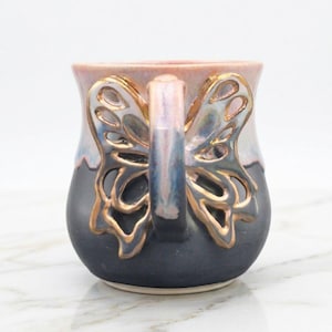 May include: A ceramic mug with a butterfly wing handle. The mug features a dark blue base, a light pink rim, and a gold-coloured butterfly wing handle. The handle has an intricate open design.