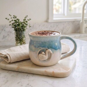 Può includere: Tazza in ceramica fatta a mano con smalto blu e bianco, riempita con una bevanda al cioccolato e guarnita con scaglie di cioccolato. La tazza presenta un disegno a rilievo di cigno e un manico curvo. La tazza è su un tagliere di marmo.