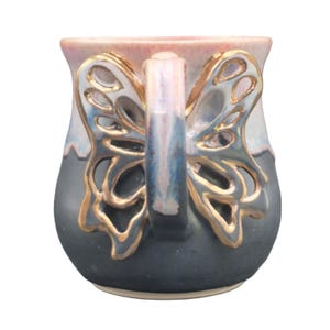 May include: A ceramic mug with a butterfly wing handle. The mug has a colour gradient of blue, grey, and pink. The butterfly wings are outlined in gold. This decorative mug is a unique item for the home.