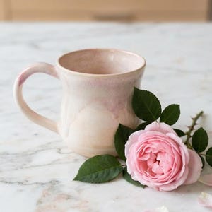 May include: A handmade ceramic mug with a light pink glaze. A pink rose with green leaves rests beside the mug. The mug has a curved handle and a wide opening. The mug is on a marble surface.